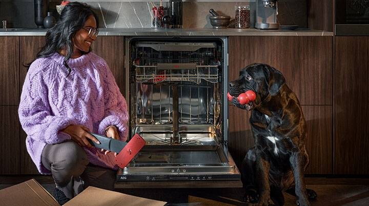 Woman unpacking spare parts for dishwashing, while smiling at a black dog with a toy in its mouth